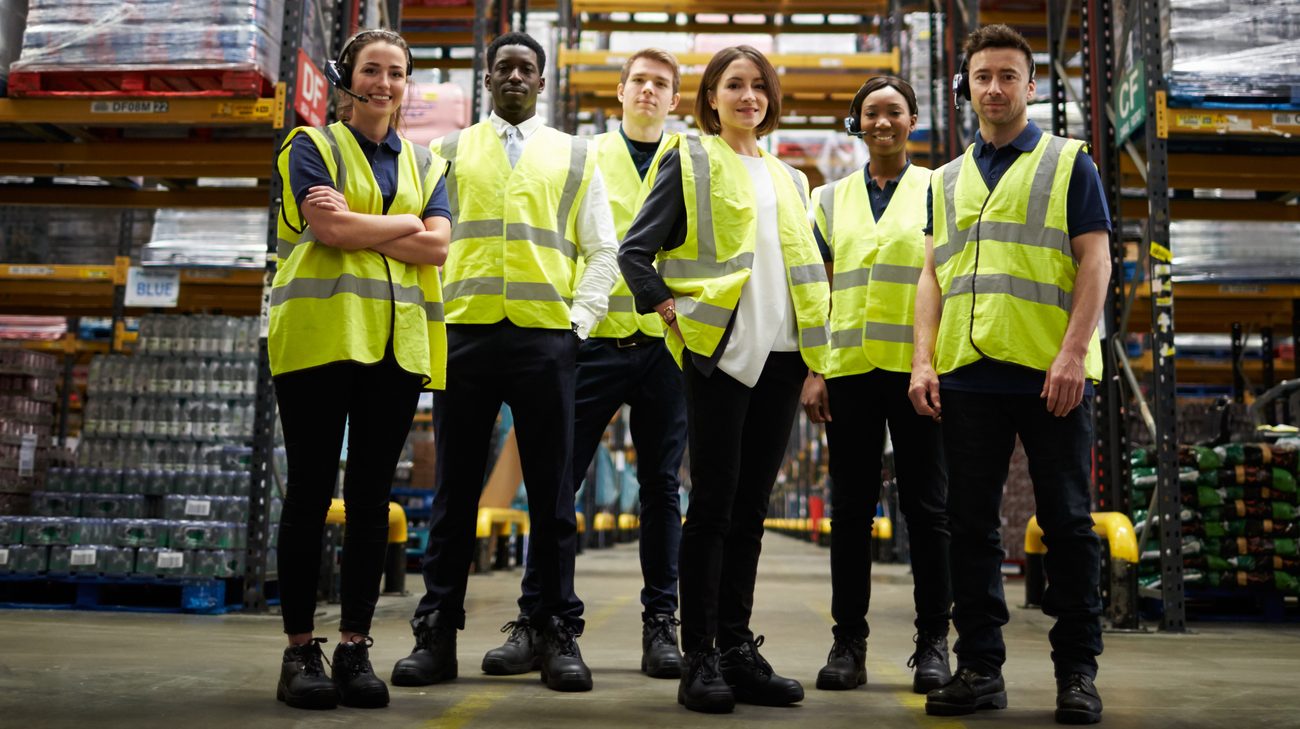 Six warehouse associates in hi-vis vests standing together in an aisle of stacked drinks pallets