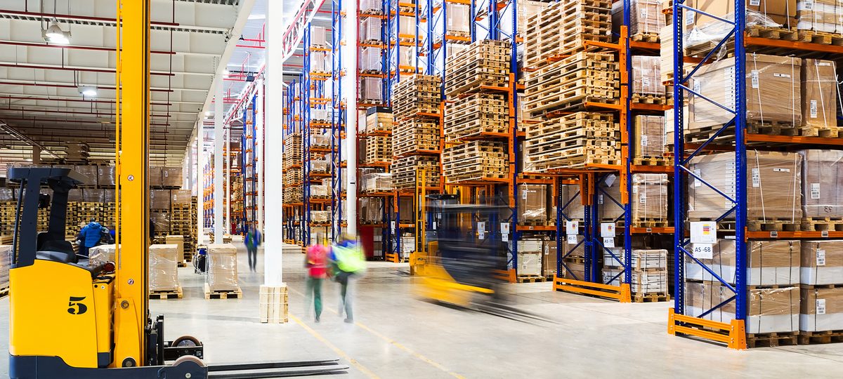 A yellow reach truck parked in a high-bay warehouse aisle lined with loaded pallet racking under bright lights