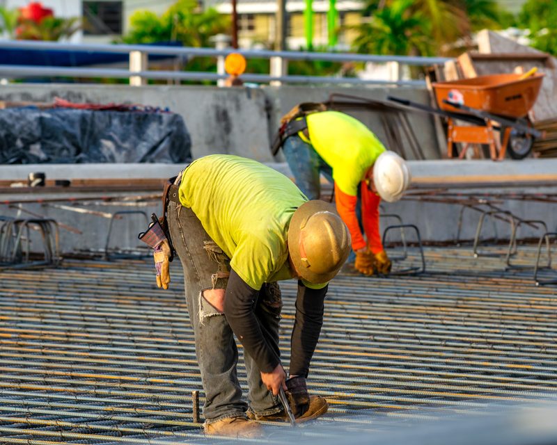 Two construction workers in hi-vis shirts tying rebar on a concrete slab under South Florida palms
