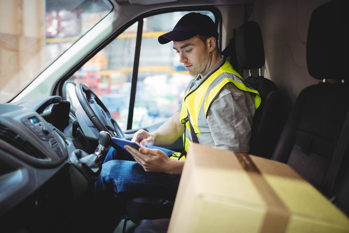 A delivery driver in a hi-vis vest checking a tablet at the wheel of his van beside a stacked cardboard box