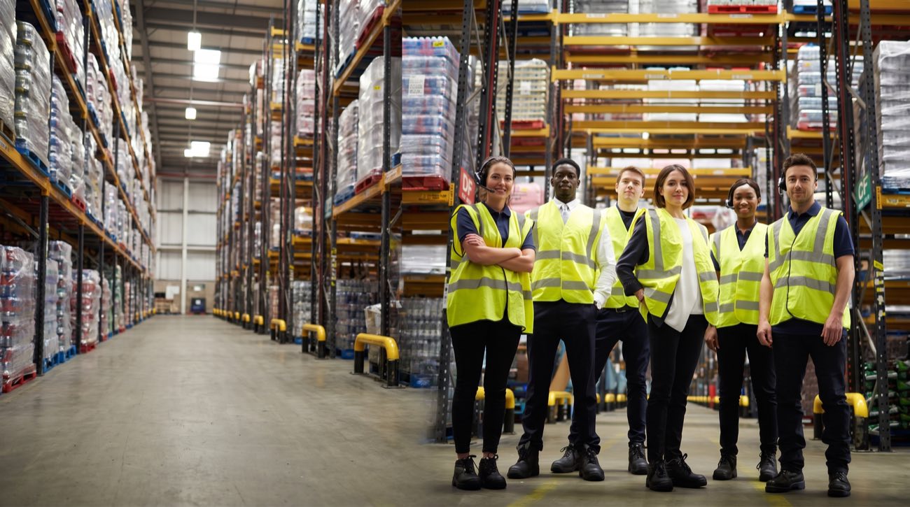 Six Support Staff Solutions workers in hi-vis vests standing together in the aisle of a stocked distribution centre
